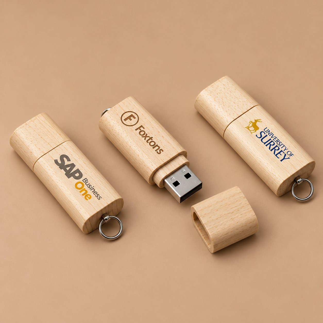 Three wooden USB drives showing SAP Business One, Foxtons and University of Surrey logos to illustrate how different marks read at real engraving size.
