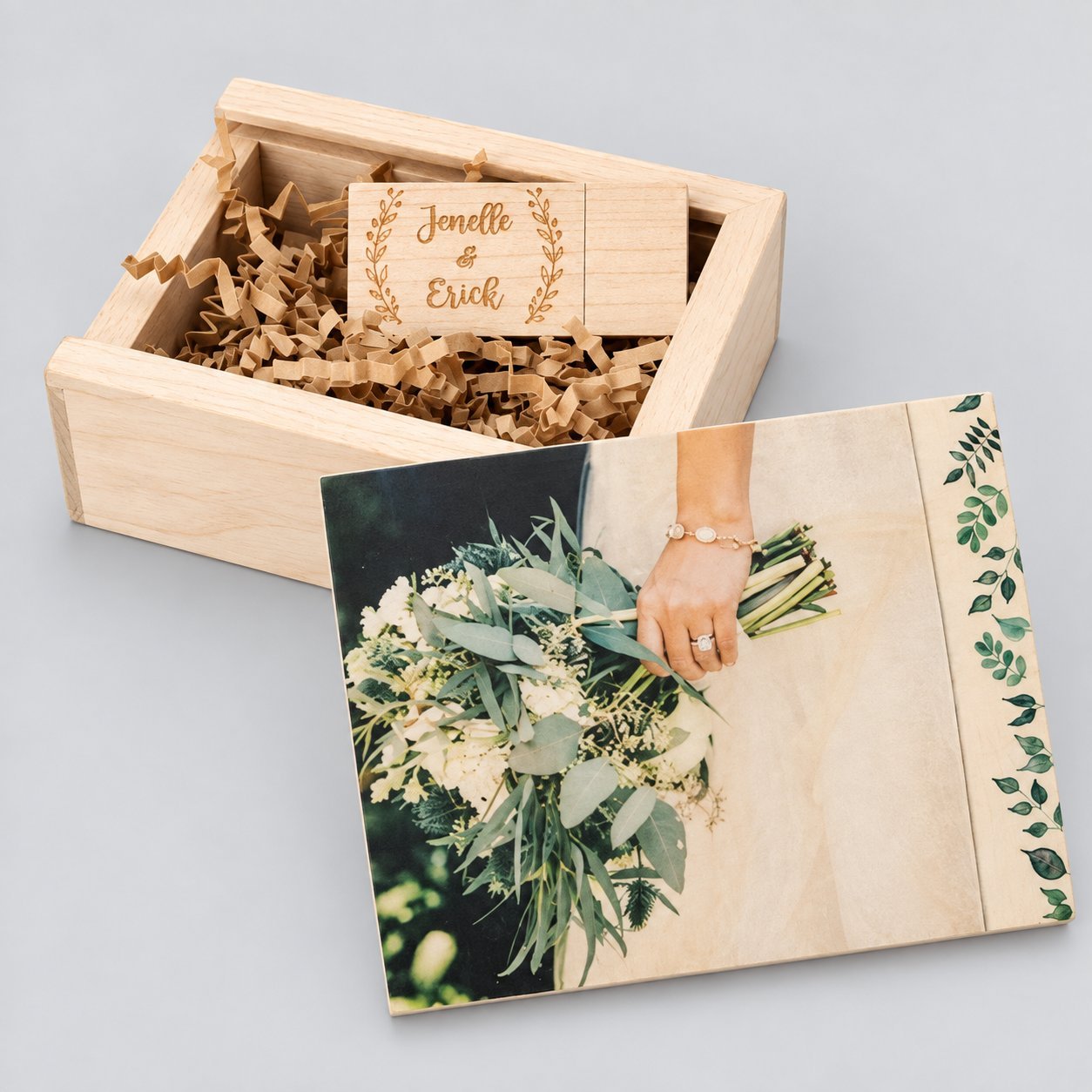 Open light-maple wooden USB gift box showing crinkled paper fill, a custom maple USB drive laser-engraved with 'Jenelle & Erich' inside the lid, and a sample 4x6 wedding photo print of a bride holding a eucalyptus bouquet in the base — illustrating how a print stack and a USB fit in a small wooden box.