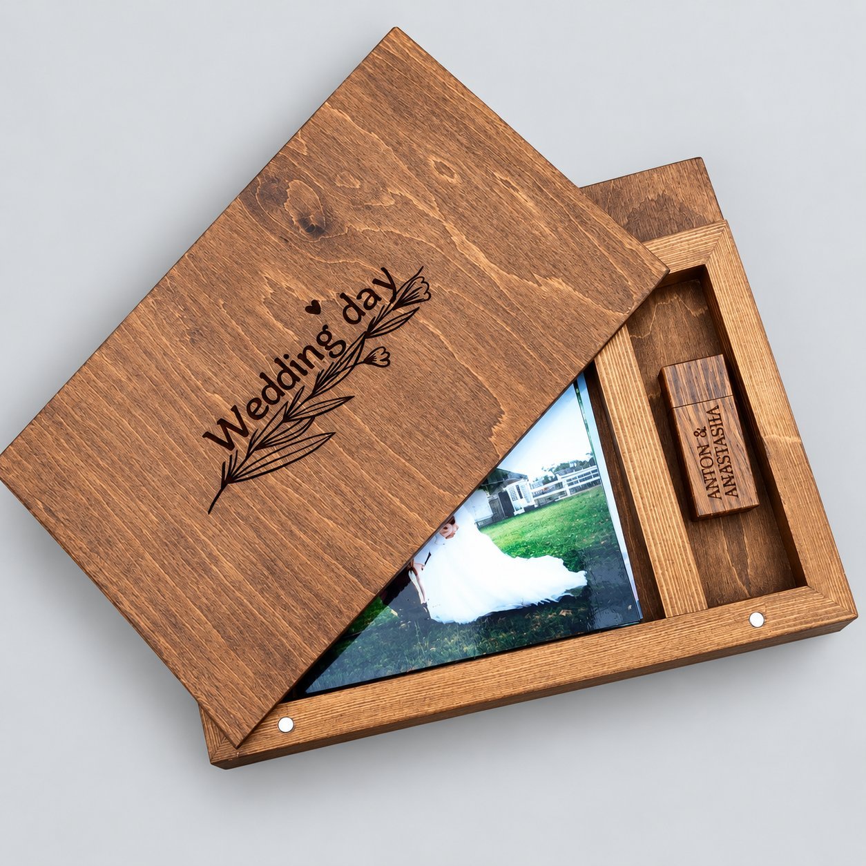 Light-oak wooden USB wedding gift box with its engraved sliding lid reading 'Wedding day' with a leafy flourish, partially pulled back to reveal a sample wedding photo print of a bride's dress and a small custom USB stick engraved 'ANTON & ANASTASIA' nested in a dedicated slot — a finished wedding-photographer sample configuration.