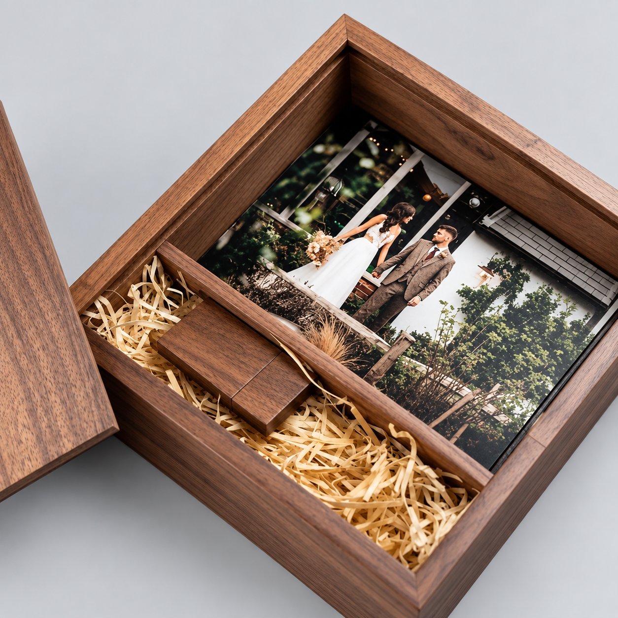 Open dark walnut wooden USB gift box with a divided die-cut foam insert: the upper compartment holds a 4x6 wedding photo print of a couple outdoors, and the lower compartment holds a custom engraved wooden USB drive nested in crinkled paper fill — showing how to keep the USB and photos in separate cavities.