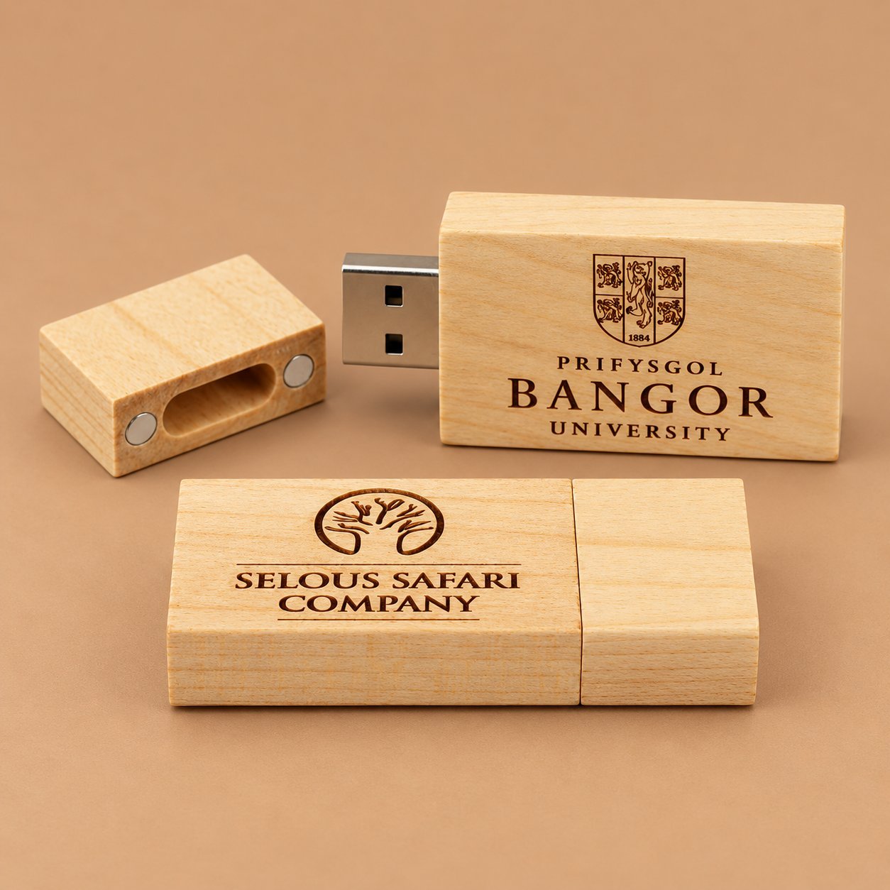 Two wooden USB drives engraved with distinct institutional marks: Bangor University with its 1884 crest, and Selous Safari Company with a deer antler logo, showing how different marks sit on similar wood bodies.