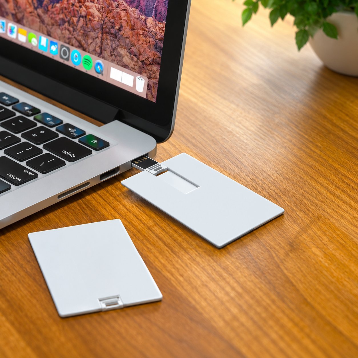Bir dizüstü bilgisayarın yanındaki ahşap bir masada, biri USB portuna takılı iki ince beyaz plastik kredi kartı tarzı USB sürücü; basılı pazarlama el ilanları için kullanılan ince plastik kart form faktörünü göstermektedir.